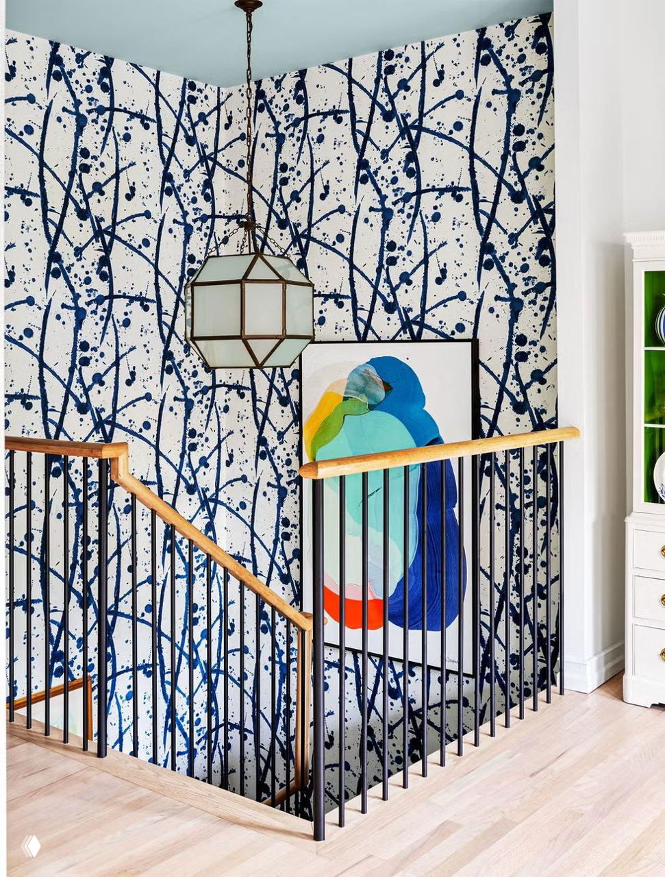 Лестничный пролет с обоями в абстрактные тёмно‑синие кляксы, деревянные поручни, чёрные металлические балясины и крупная картина у выхода вниз.