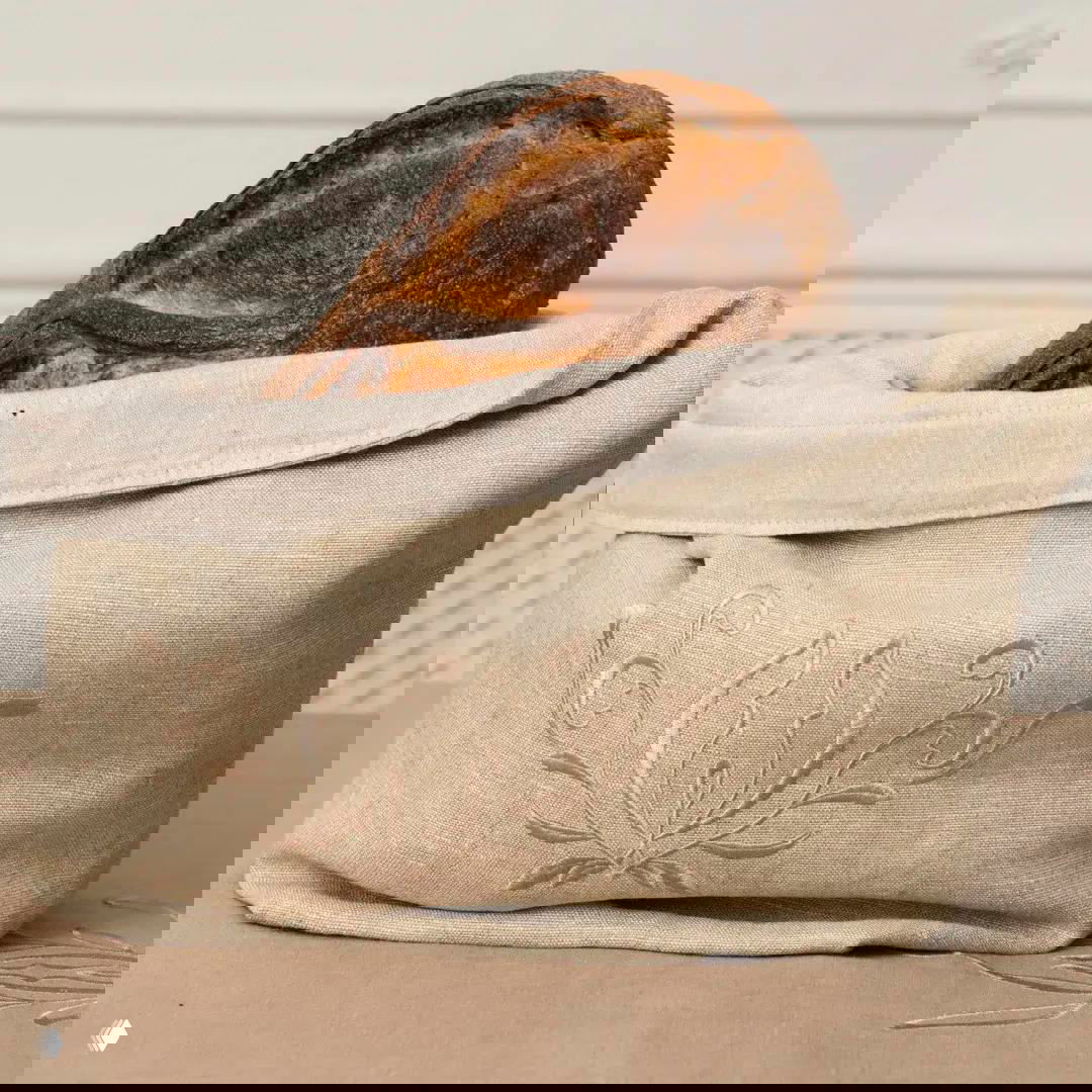 Текстильный хлебный мешок из льна с тонкой бежевой вышивкой, в котором аккуратно хранится буханка домашнего хлеба.