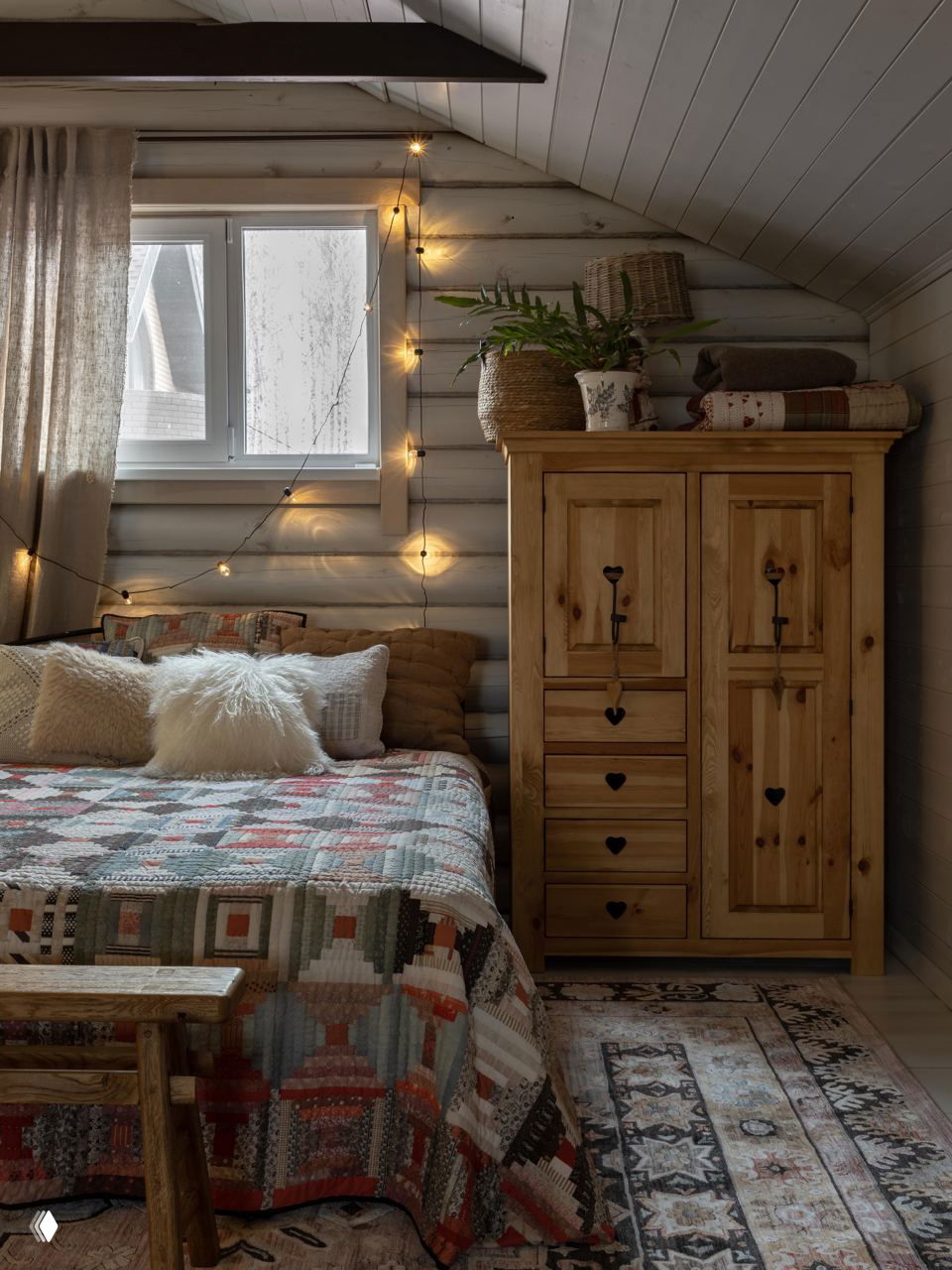 Мансардная спальня с деревянным шкафом, лоскутным покрывалом на кровати, гирляндой у окна и уютными декоративными подушками.