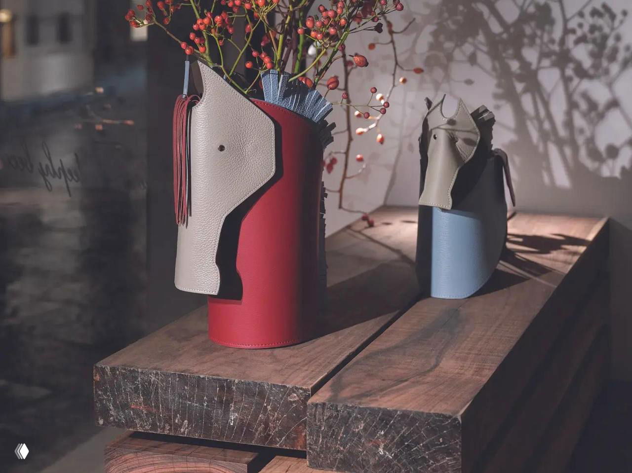 Крупный план двух кожаных аксессуаров Nuvola: высокий красный цилиндр‑вазон с элементами в виде головы лошади и голубой кожаный предмет‑подставка.