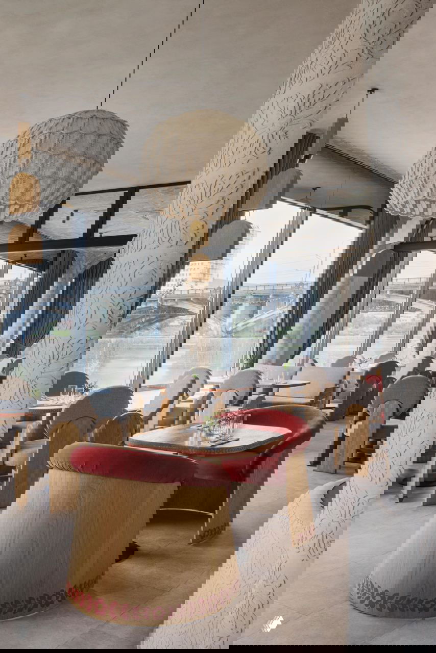 Общий вид зала с видом на мост и реку: дубовые столы и стулья, красные бархатные сиденья и крупные плетёные купольные светильники под потолком.