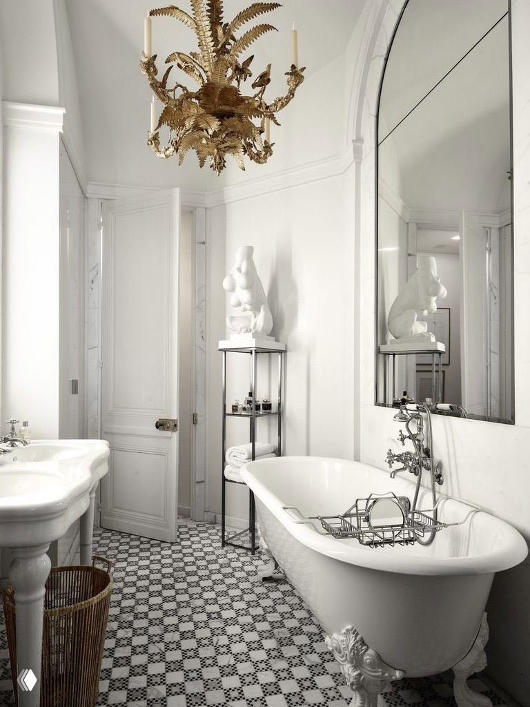 Классическая белая ванная с отдельностоящей чугунной ванной, декоративной позолоченной люстрой и узорчатой напольной плиткой; винтажные элементы в светлом интерьере.