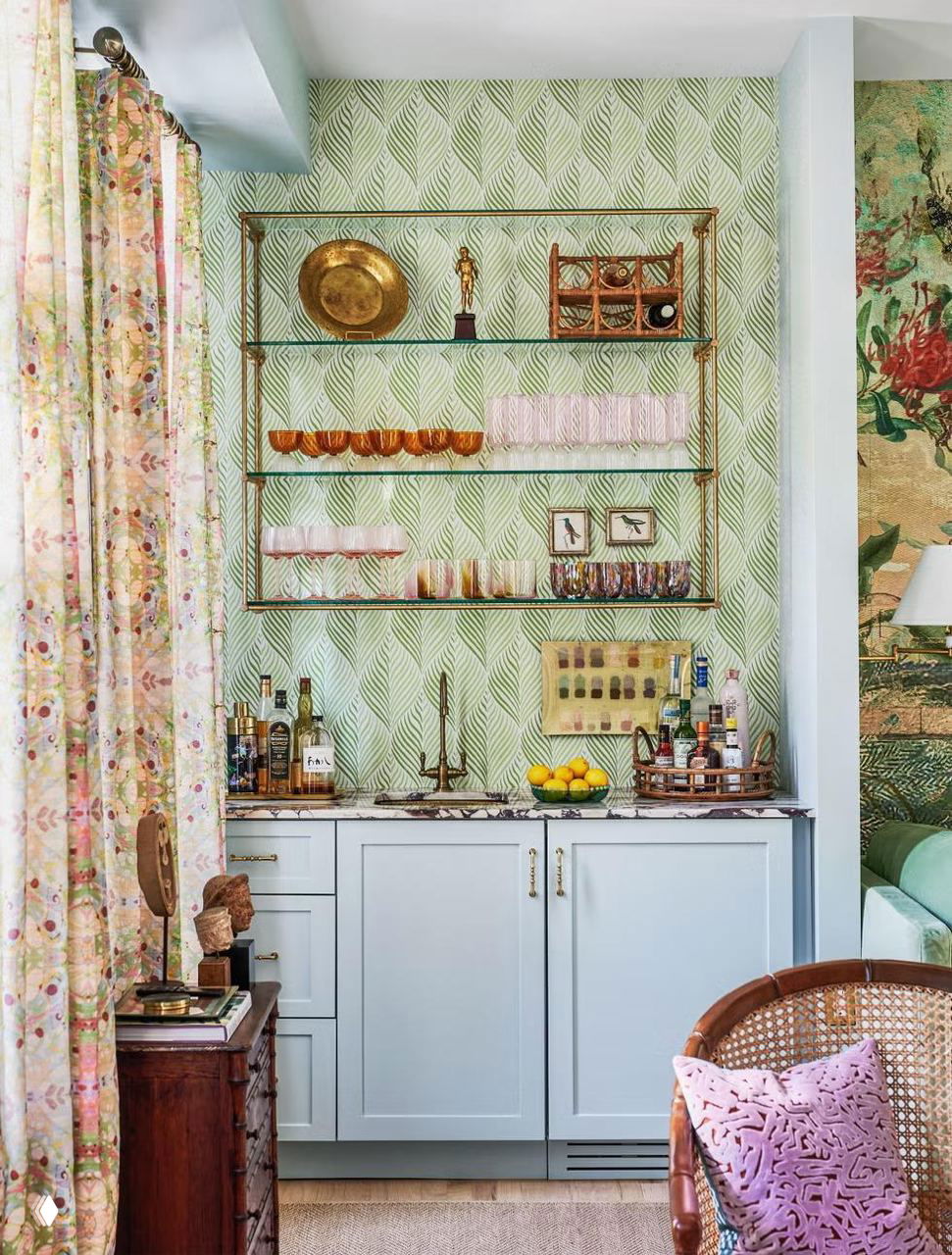 Небольшая барная зона с зелёными обоями в листовой узор, золотая металлическая полка, пастельно‑голубые шкафчики и предметы бара на мраморной столешнице.