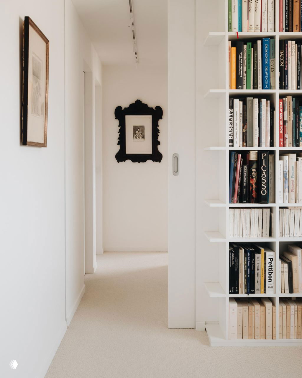Узкий белый коридор с встроенной книжной полкой справа и картинами на стене; организованная книжная коллекция и светлый пол создают уютное пространство.