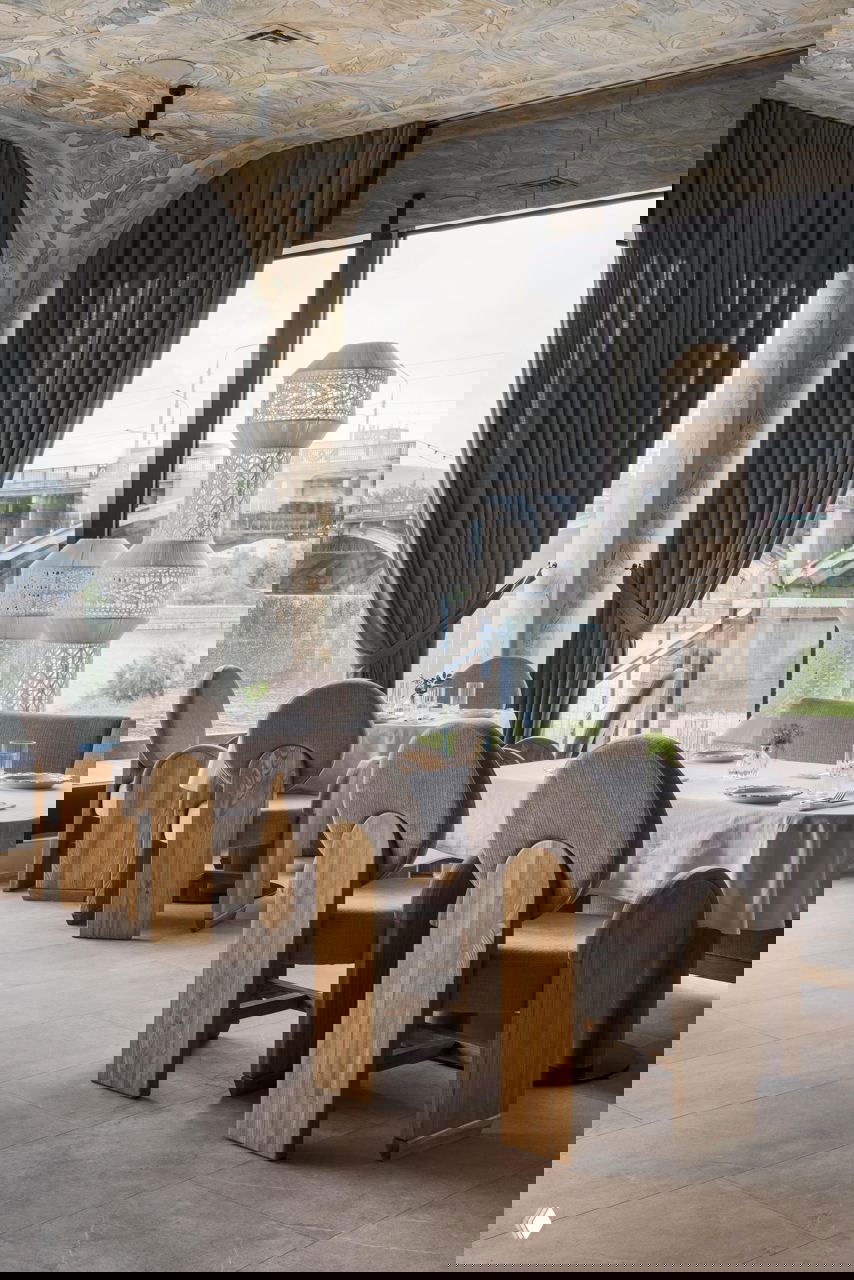 Столы у больших окон: сервировка на белых скатертях, округлые спинки стульев и декоративные арочные колонны с растительным орнаментом на фоне реки.