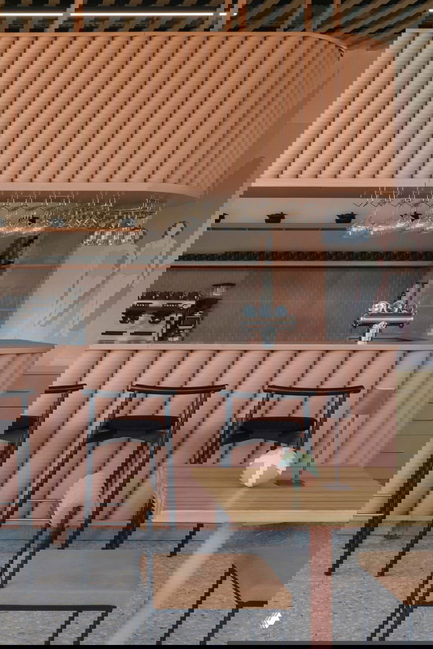 Ближний план барной стойки с вертикальными цилиндрическими панелями в коралловом тоне, высокие черные барные стулья и подвешенные бокалы над стойкой.