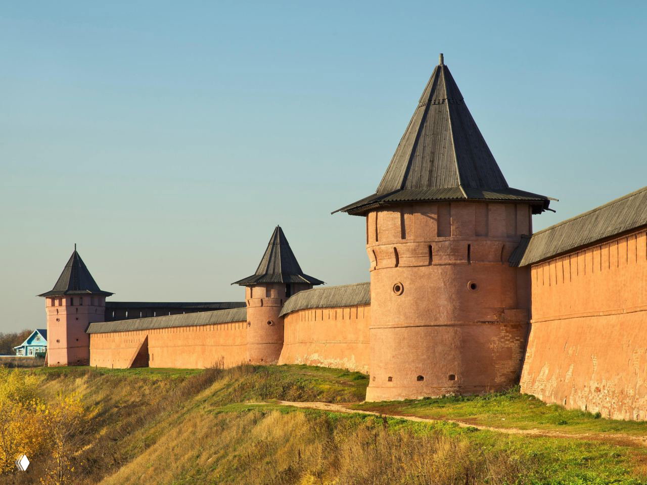 Части крепостной стены и башни Суздальского кремля на фоне светлого неба и прибрежного склона, видимый защитный вал и дорожка вдоль стены.