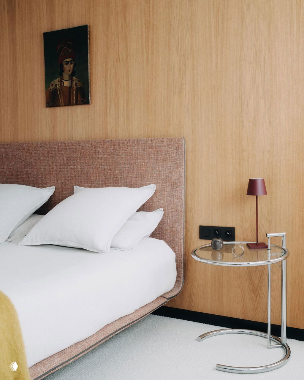 Спальня с мягкой обитой кроватью и белыми текстильными элементами на фоне деревянной стеновой панели; круглый стеклянный прикроватный столик и небольшая лампа.