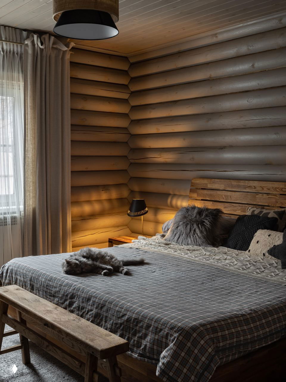 Главная спальня с оцилиндрованной бревенчатой стеной, большой кроватью с мягкими покрывалами и тёплым боковым освещением у прикроватной тумбы.