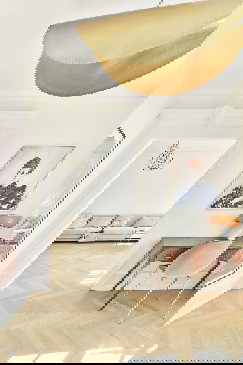 Крупный план золотистой полукруглой люстры Gervasoni в просторной гостиной с лепниной, арт-объектами и теплыми нейтральными акцентами.