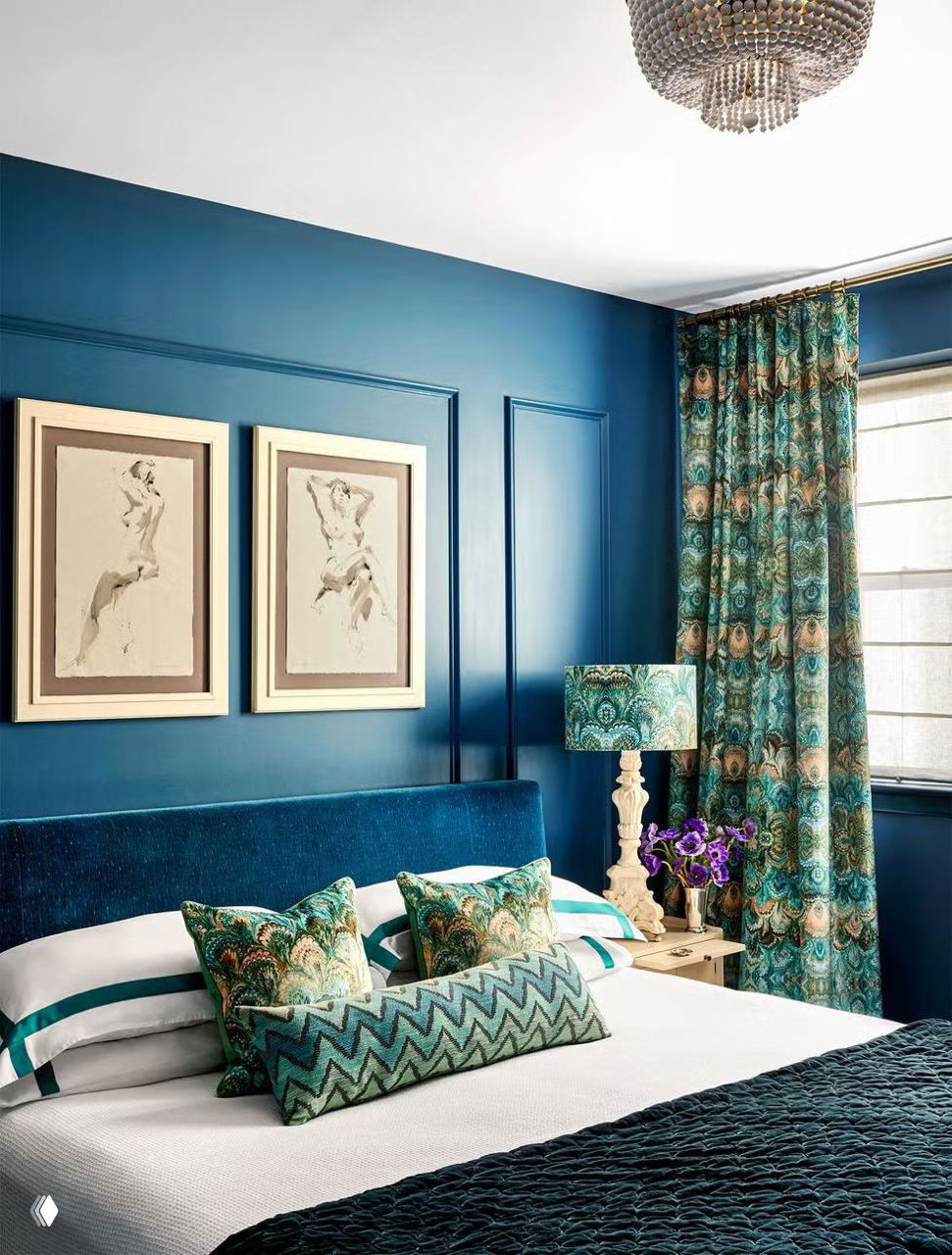 Спальня в насыщенных бирюзово‑синих тонах: глубокие синие стены, бархатное изголовье, декоративные подушки и шторы с пейзажным узором, люстра.