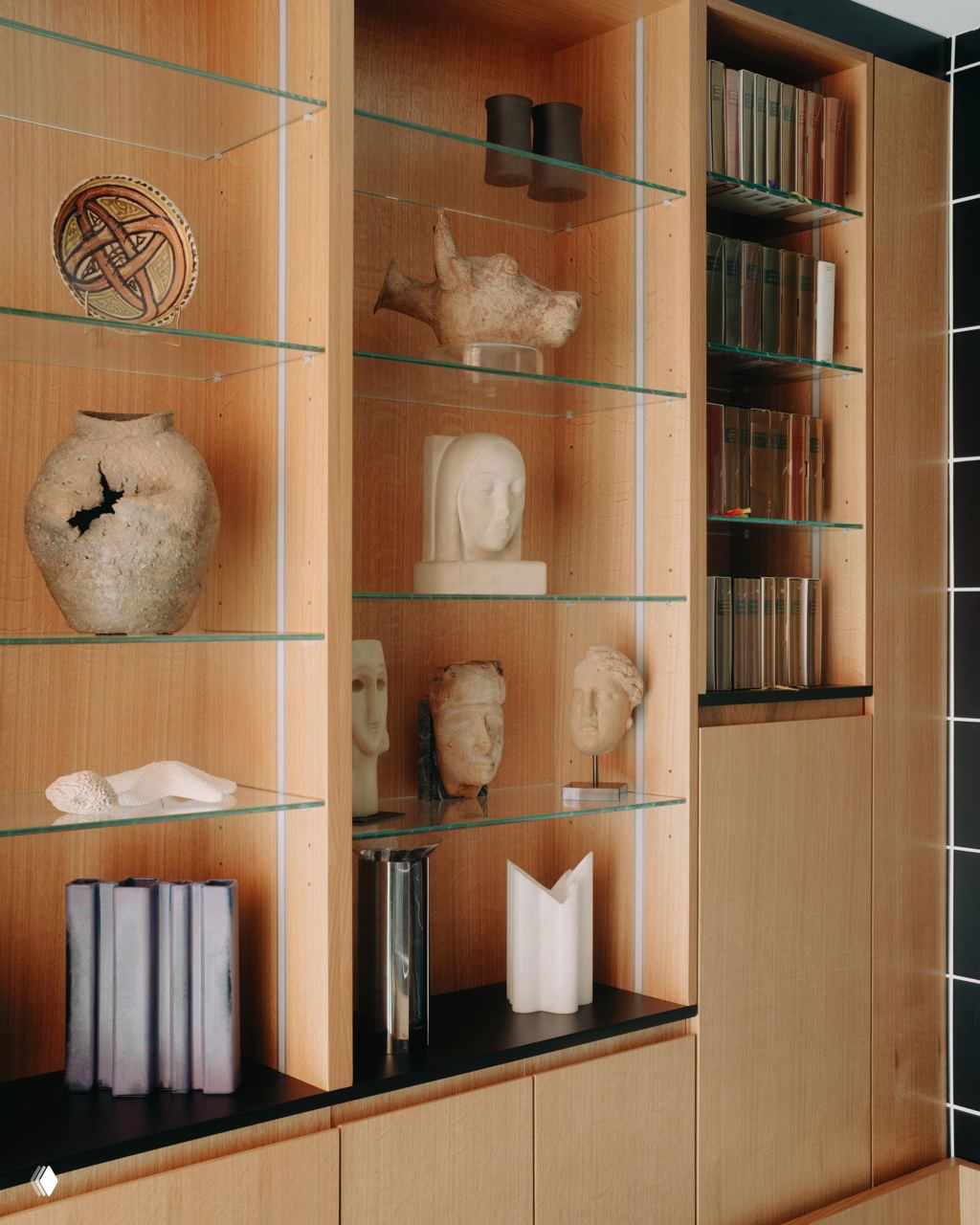 Витрина с деревянными полками и стеклянными вставками, на которых выставлены керамические сосуды, скульптурные головы и декоративные книги — коллекционный декор.