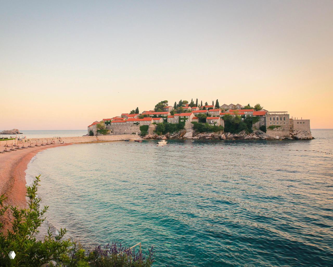 Спокойный прибрежный пейзаж с небольшим островом и домами с красными крышами, пляж и гладкая морская поверхность при тёплом вечернем свете.