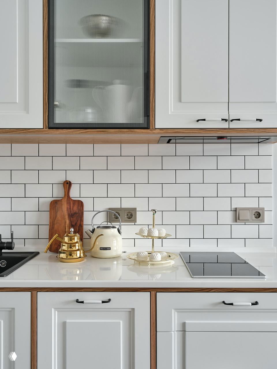 Крупный план кухонной рабочей зоны: белая столешница, деревянные вставки шкафов и золотистые аксессуары на фоне белой плитки.
