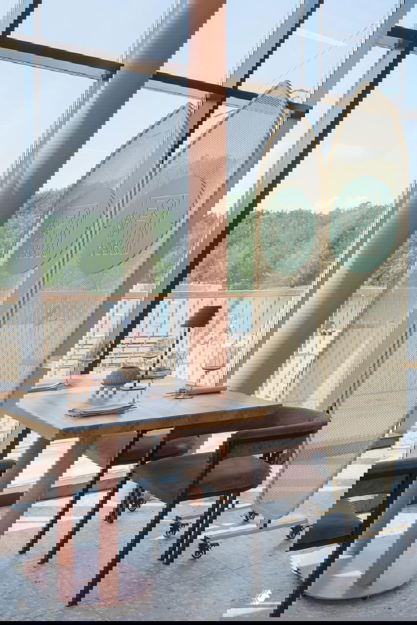 Обеденная зона у панорамных окон: стол с розовой опорой, кресла и перегородки-«серфы» с логотипом ресторана; вид на пляж и деревья за стеклом.