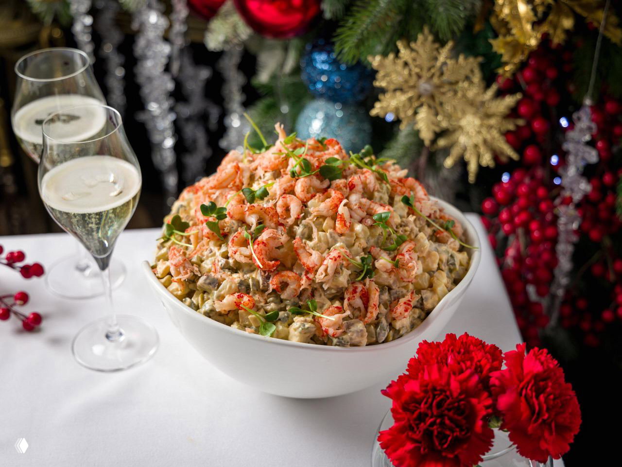 Праздничный салат оливье с креветками или раковыми шейками в белой миске на фоне новогодних украшений и бокалов шампанского