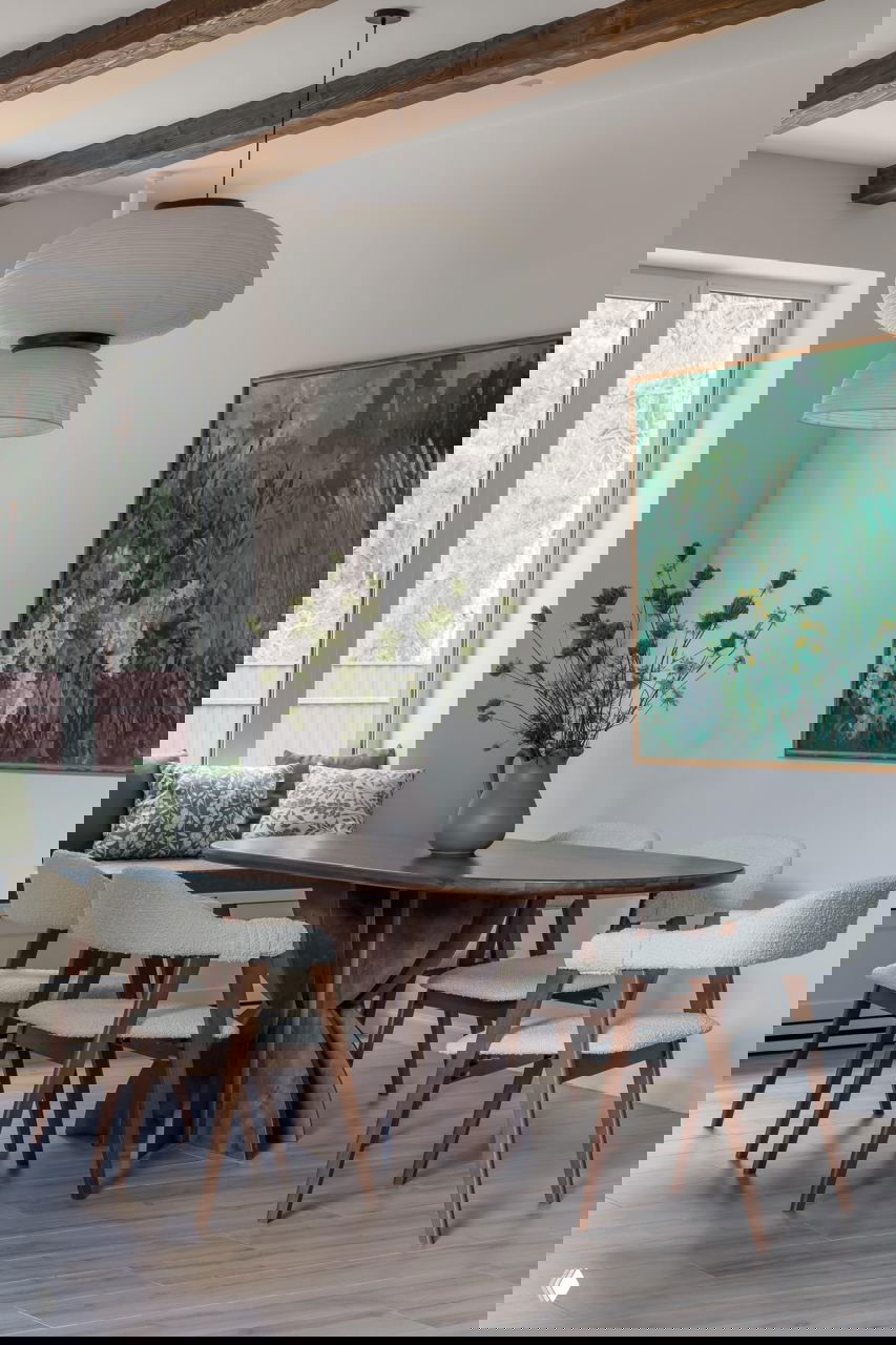 Столовая зона с круглым деревянным столом, светлыми обитыми креслами и большой живописной картиной в зелёной палитре; потолочные балки добавляют текстуру интерьеру.