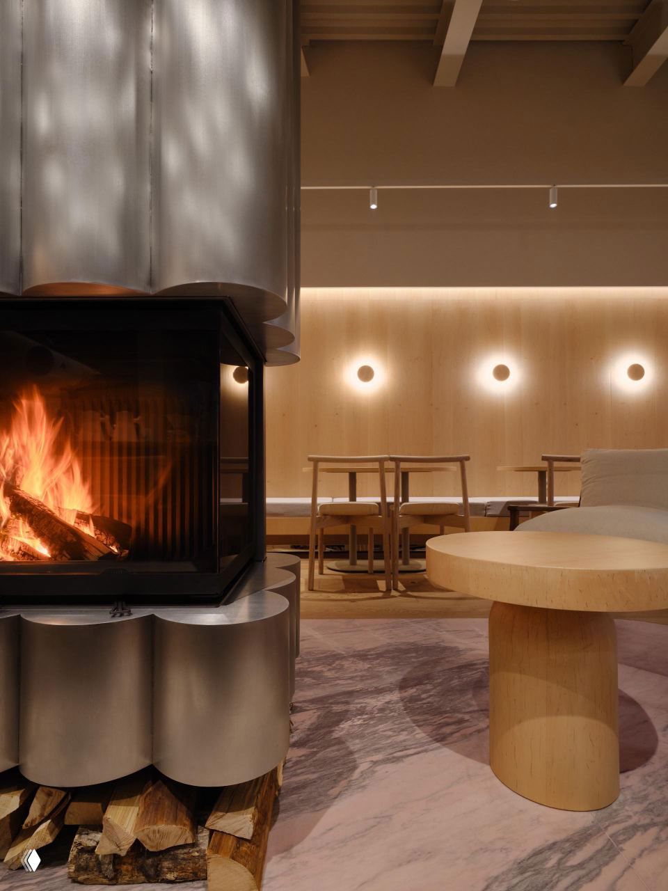 Камин из стальных цилиндров с горящим дровяным огнём, деревянная мебель и мягкое тёплое освещение создают атмосферу загородного дома внутри кафе.