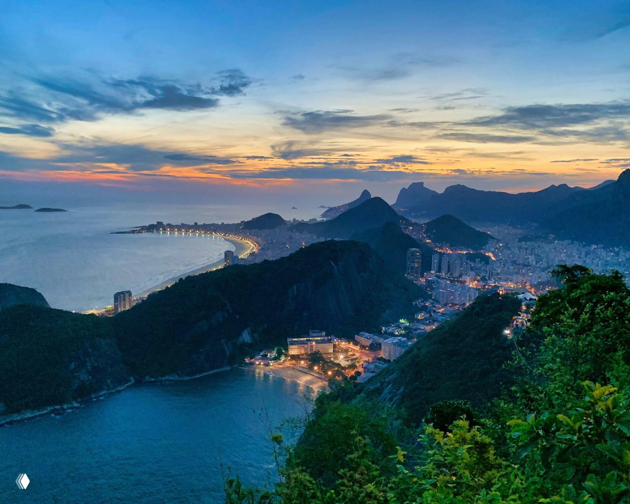 Вид с высоты на извилистую береговую линию, залив и городской пейзаж с огнями вечером; горы, леса и тёмное море создают контрастную сцену.