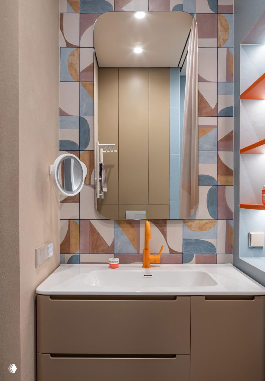 Ванная комната и раковина на фоне декоративной графичной плитки в пастельных тонах; оранжевый смеситель и организованные ниши придают интерьеру характер.
