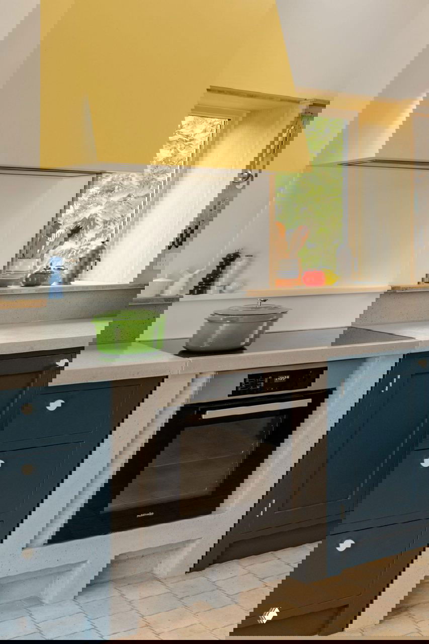 Кухонная плита и духовка встроены в бетонную стойку, над ними ярко-жёллый кухонный козырёк и зелёная кастрюля на плите.