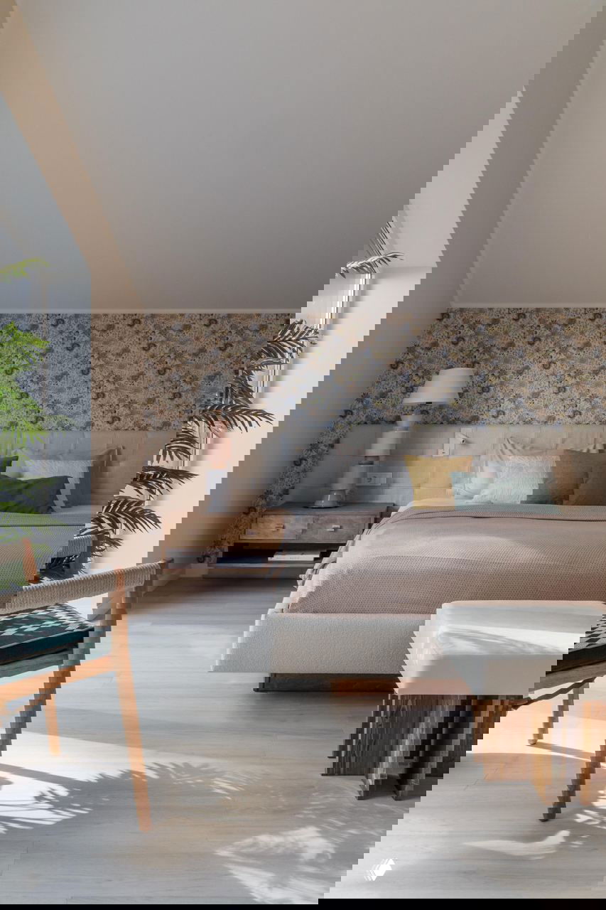 Уютная спальня в мягких бежевых тонах под скатом крыши с цветочными обоями за изголовьем и большой кроватью, солнечные лучи на светлом полу.