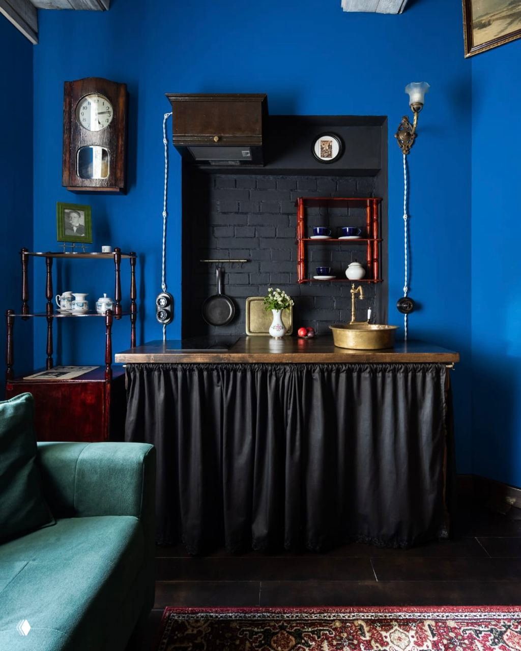 Интимная кухня с глубоко‑синими стенами и чёрной нишей над столешницей, старинные часы и открытые полки создают винтажный характер интерьера.
