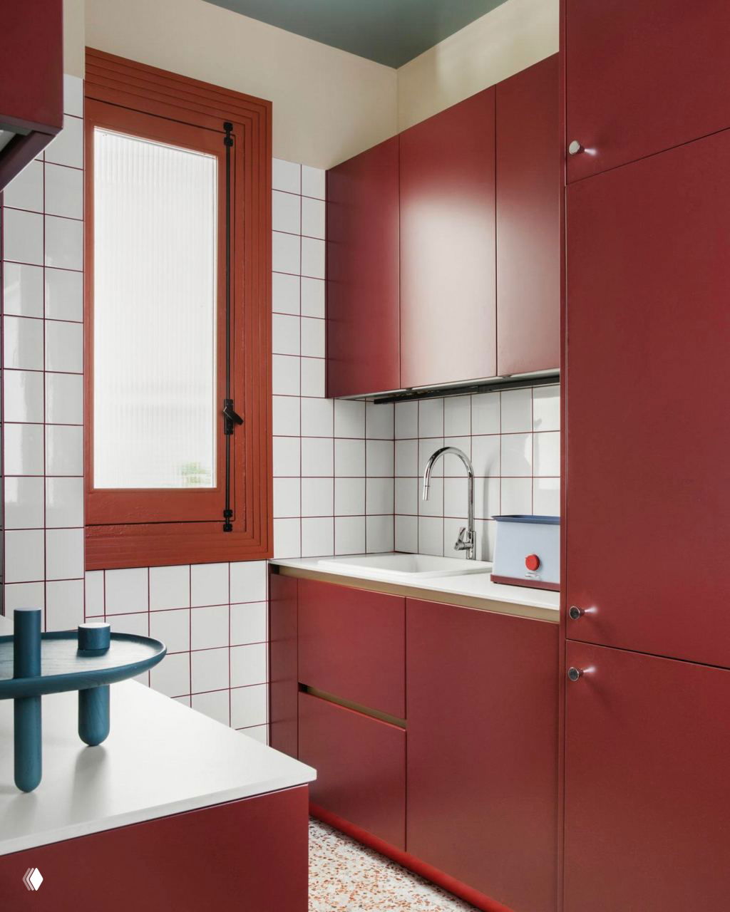 Компактная кухня с матовыми бордовыми фасадами, белой квадратной плиткой на фартуке и деревянной рамой окна коричневого цвета.