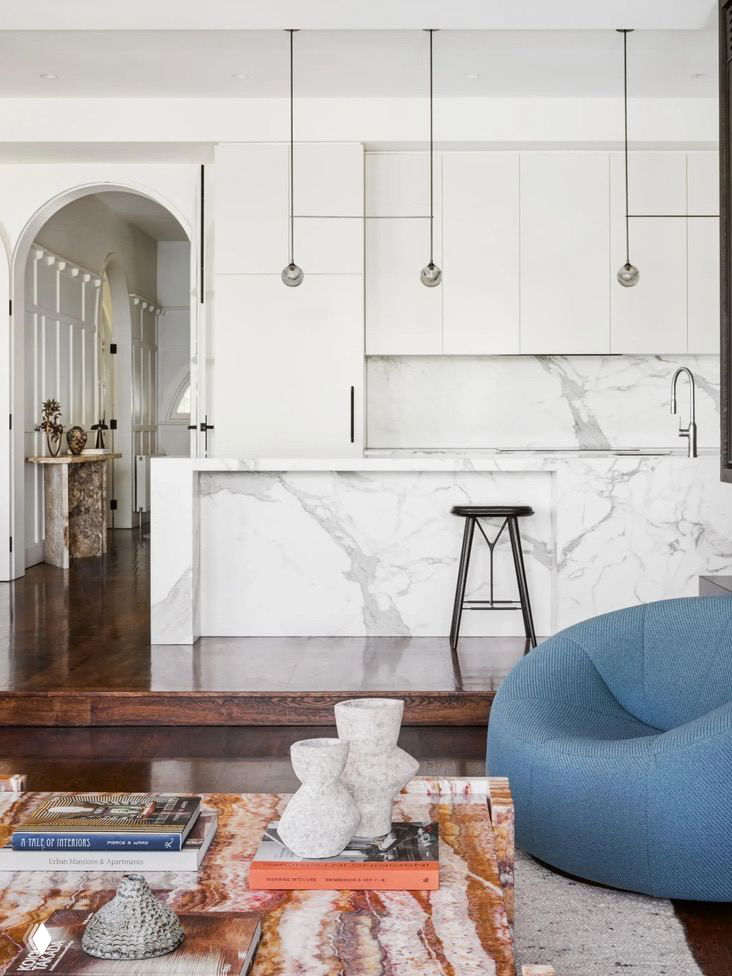 Светлая кухня в викторианском доме: белая кухонная зона с мраморной столешницей, арочный проход и сочетание классической архитектуры с минималистичной отделкой и креслом‑пуфом синего цвета.