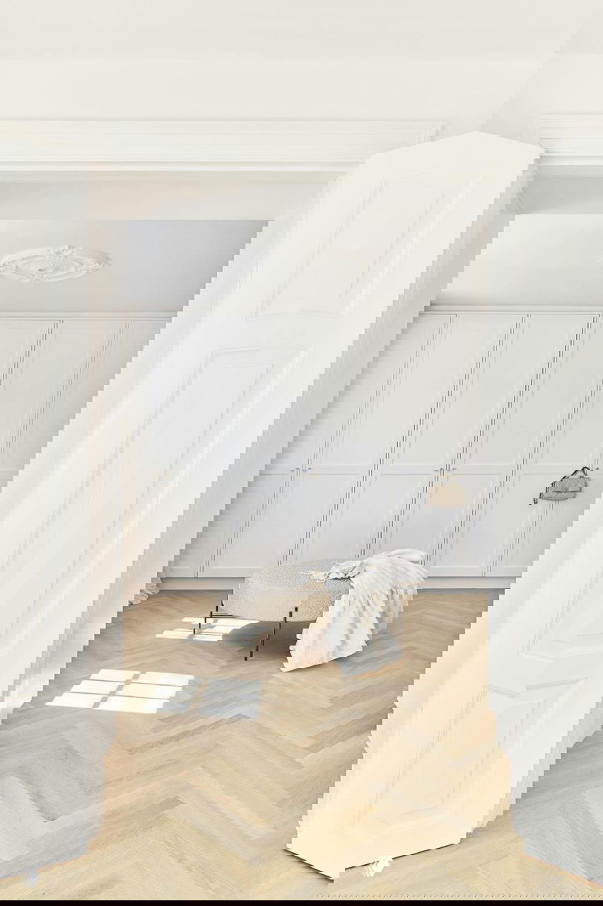 Просторная гардеробная зона с белыми встроенными шкафами, круглым пуфом и светлым паркетом — пастельная гамма и естественный дневной свет.