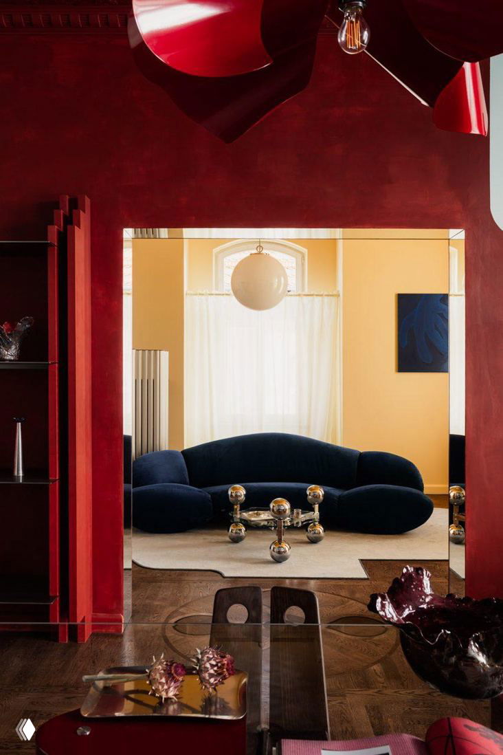 Вид через бордовую арку в комнату с синим бархатным диваном и металлическим журнальным столиком-шариками — сочетание текстур и цветов в интерьере.