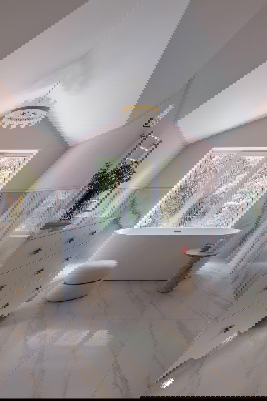 Ванная комната на мансардном уровне: отдельная свободностоящая ванна у большого окна с видом на лес, роскошная плитка с мраморным рисунком и мягкая пастельная палитра.