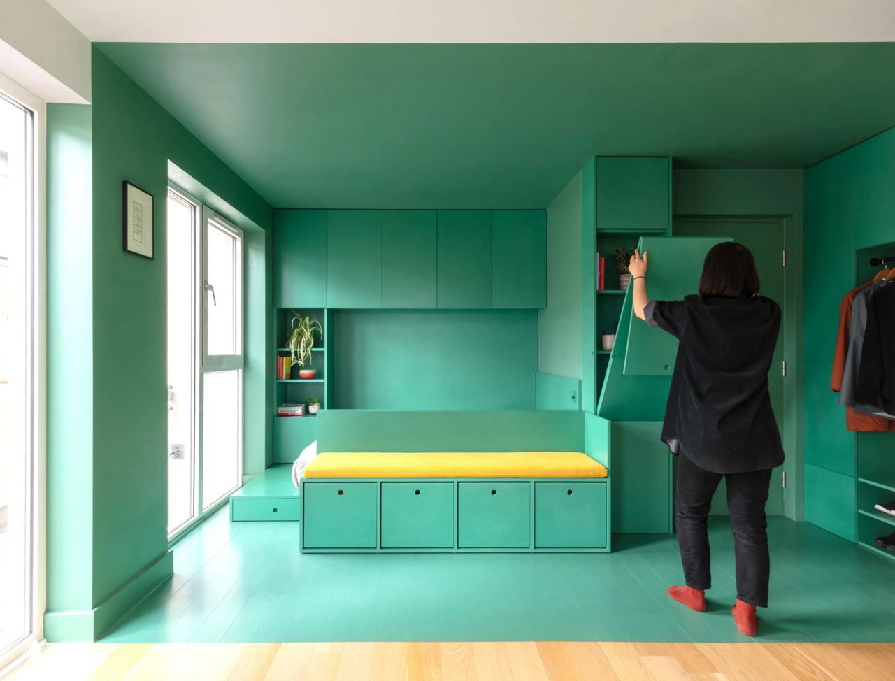 Встроенная мебель и раскладывающиеся элементы в компактной комнате: диван-кровать с жёлтой подушкой и открывающиеся фасады шкафов, человек открывает модуль