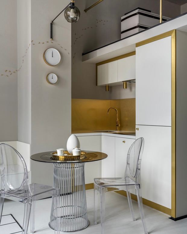 Минималистичная бело‑золотая кухня в нише с прозрачными пластиковыми стульями и круглым стеклянным столом, элегантное сочетание материалов.