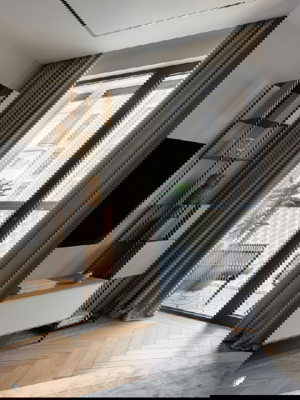 Зона гостиной и выход на балкон: высокие окна, плотные серые шторы, открытая полка с декоративными объектами и светлый паркет.