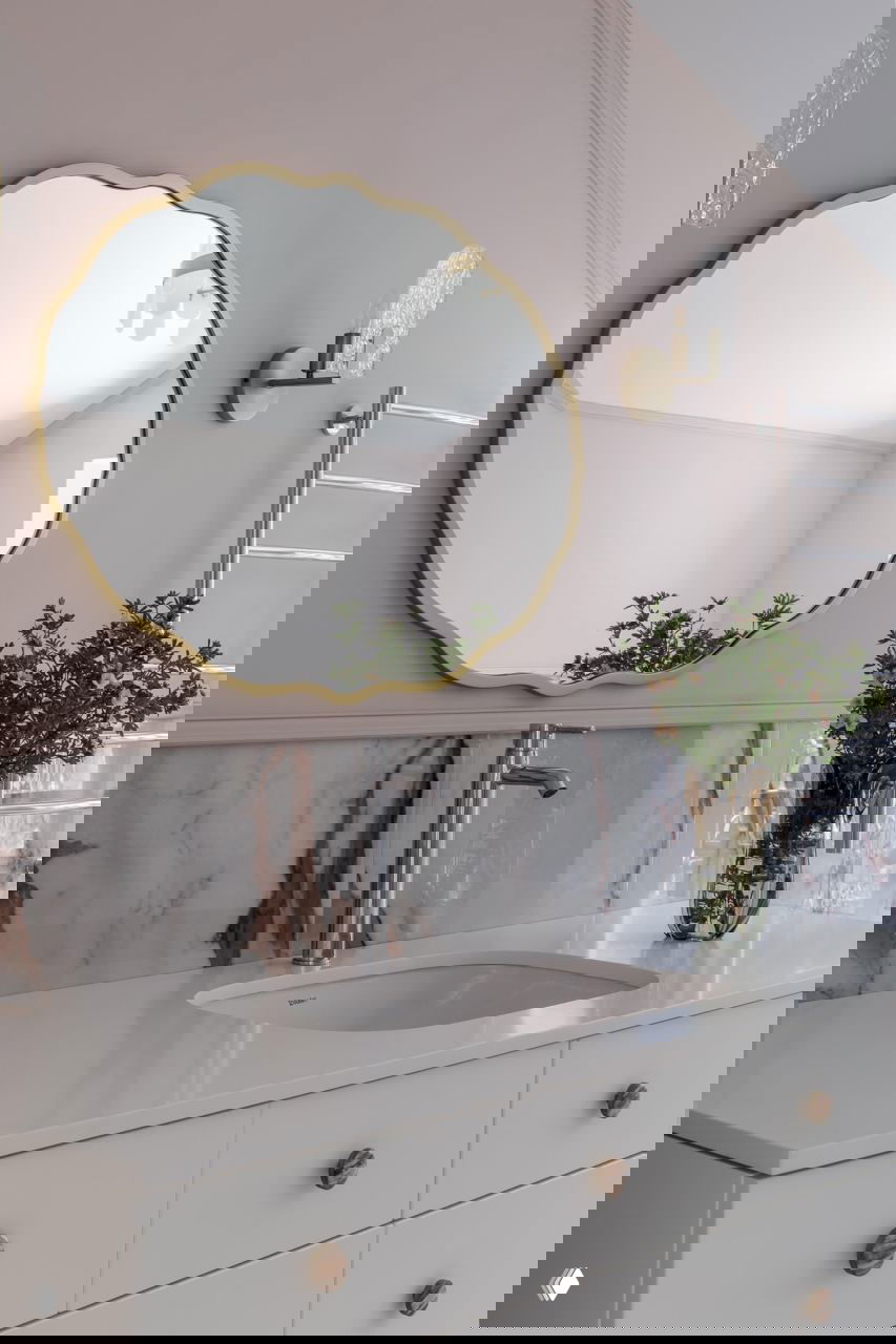 Крупный план раковины и столешницы в ванной, круглое декоративное зеркало в золотой оправе и вазочка с зелёной композицией — акцент на деталях отделки и аксессуарах.