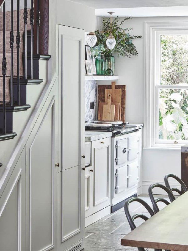 Узкая кухонная зона у лестницы с высоким встроенным шкафом до потолка, окном и обеденной зоной — использование пространства под лестницей.