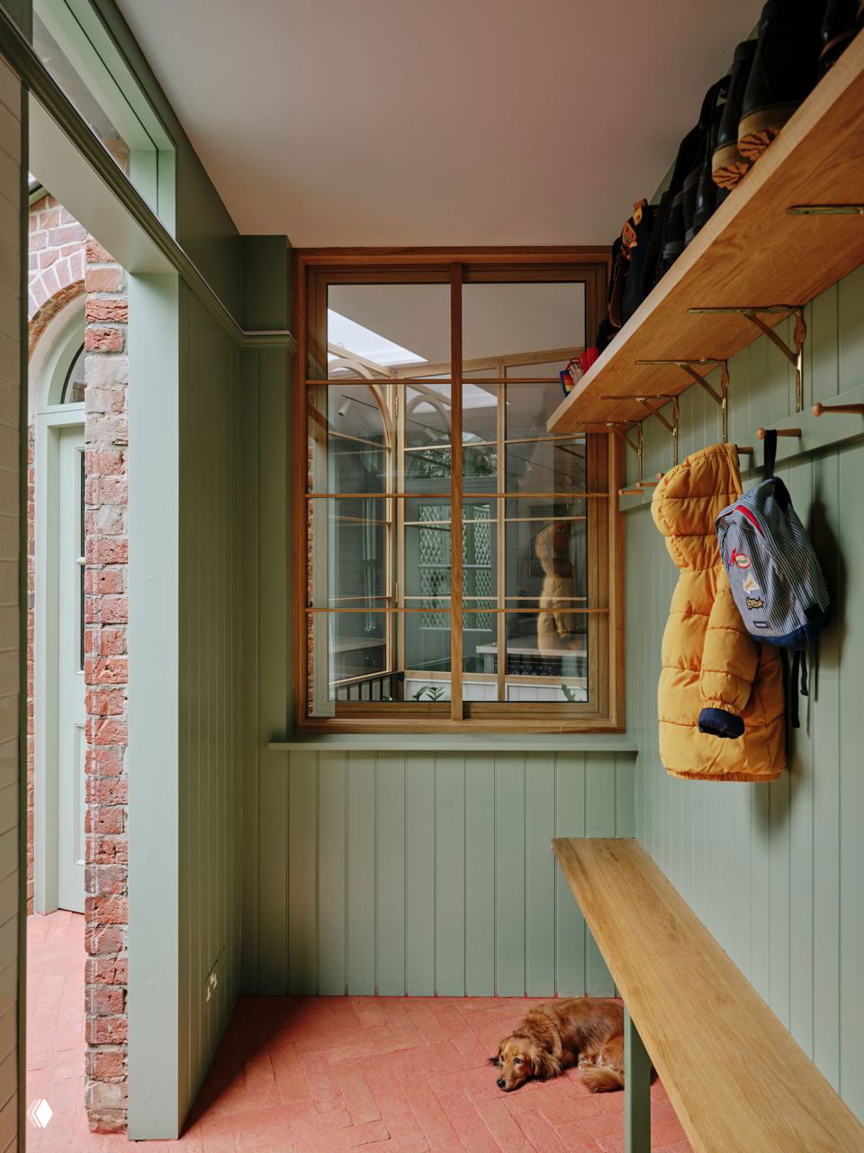 Прихожая с зелёной деревянной обшивкой, скамьёй и крючками для верхней одежды; на полу кирпичная плитка и спящий коричневый пёс у двери.