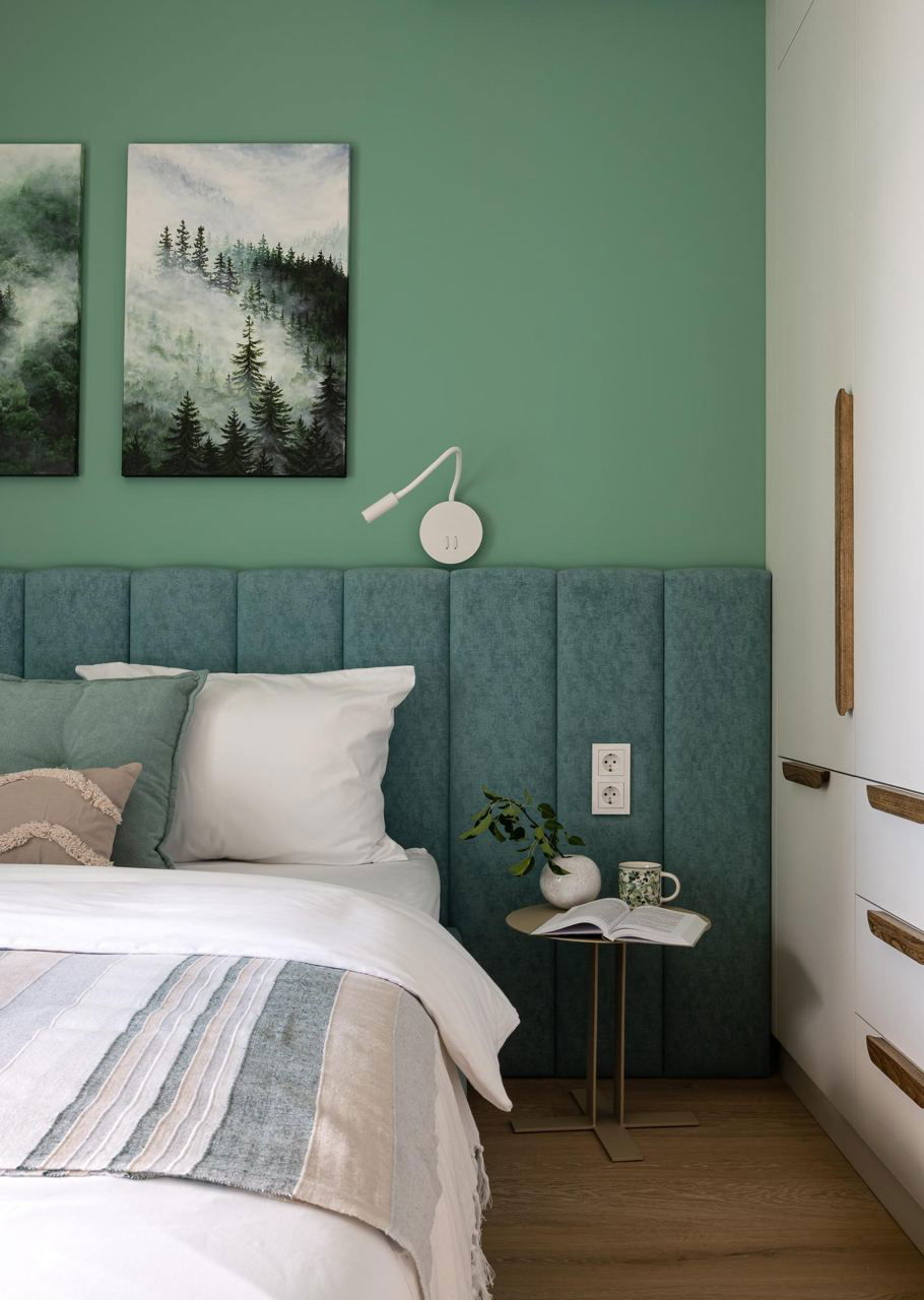 Спальня с изголовьем мягкого бирюзового оттенка, текстиль в природных тонах и пара декоративных картин с лесными пейзажами над кроватью.