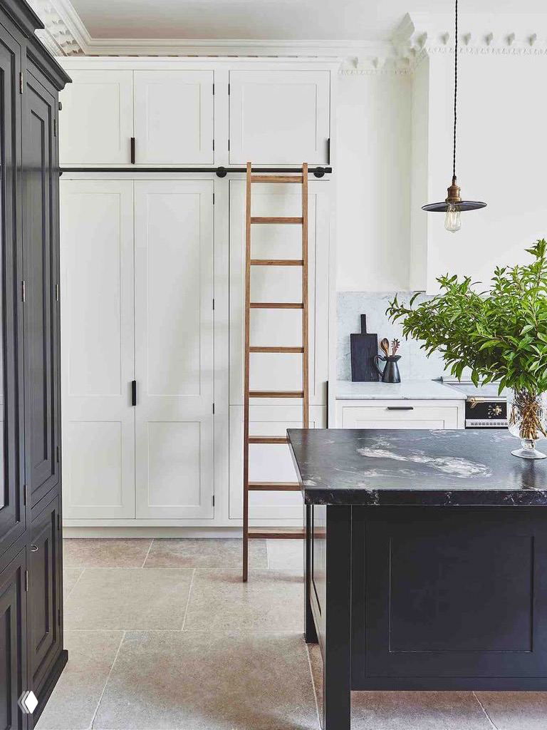 Белые высокие шкафы с передвижной лестницей и рабочий остров — решение для хранения в помещениях с высокими потолками и классическим декором.