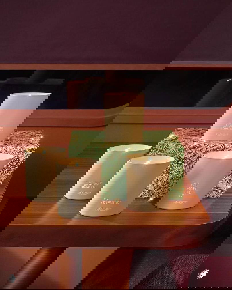 Несколько зажжённых свечей в матовых кремовых стаканах на деревянной поверхности и подарочная коробка с растительным узором