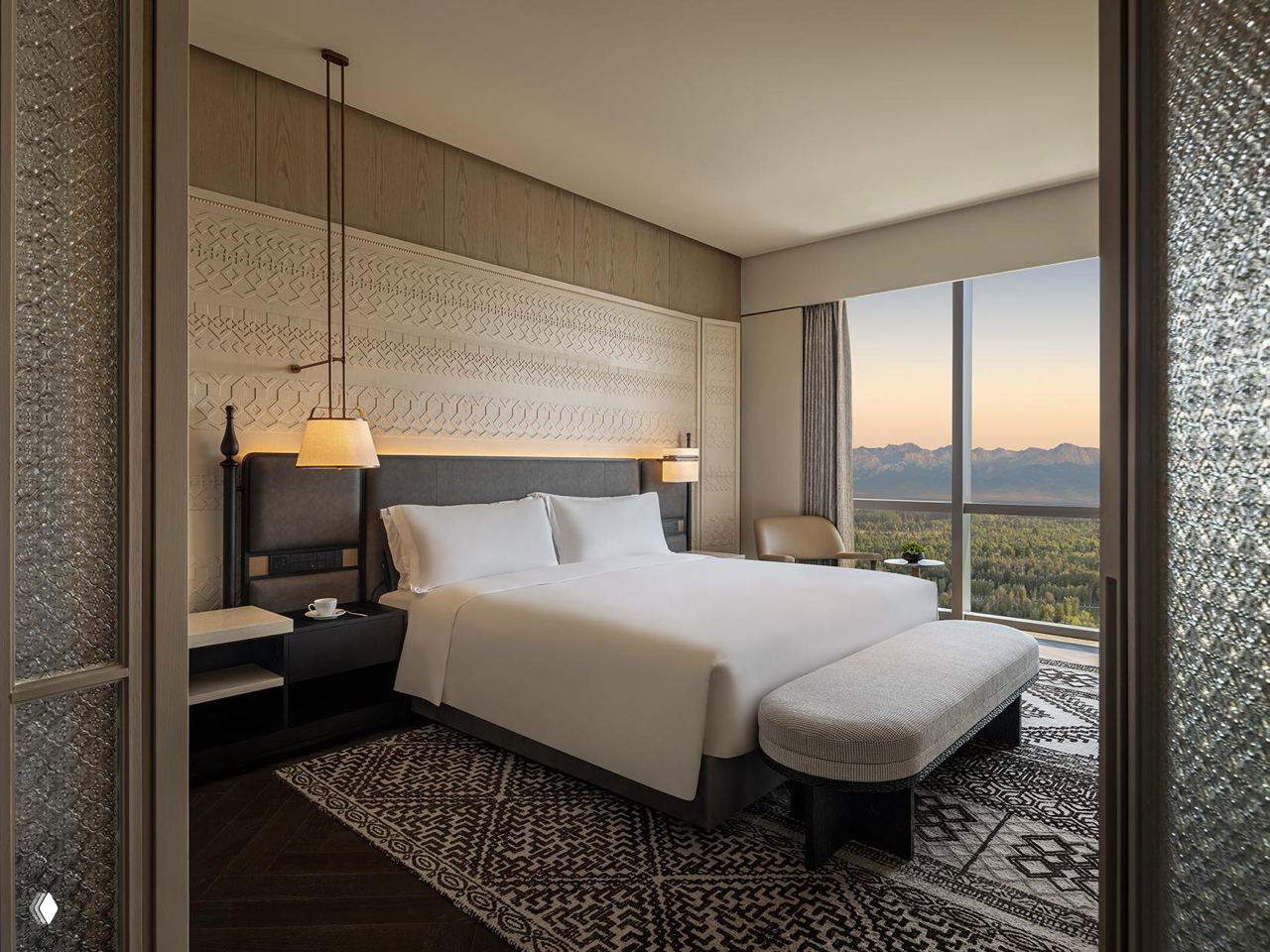 Номер отеля InterContinental: большая кровать, сдержанная отделка, резной орнамент за изголовьем и панорамное окно с видом на долину и горы Тянь‑Шаня вдалеке.
