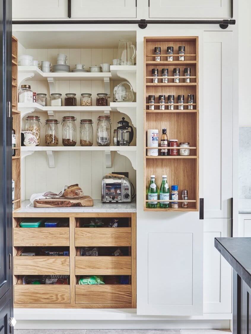 Открытая кладовка с полками и выдвижными деревянными ящиками, заполненная банками и кухонными принадлежностями — пример аккуратного зонального хранения.