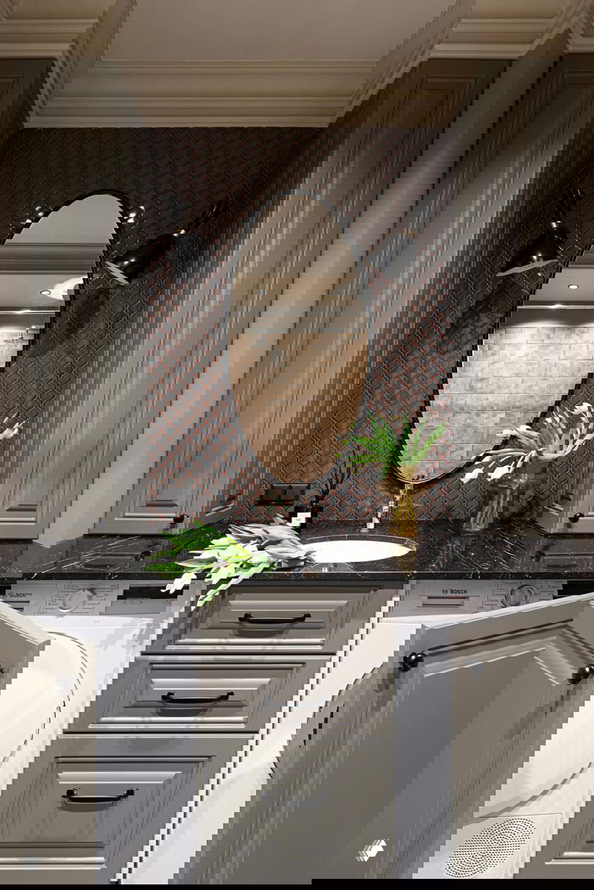 Санузел с чёрной столешницей, овальным зеркалом, встроенными шкафами и стиральной техникой, оформленный в нейтральной палитре и текстурной плитке.