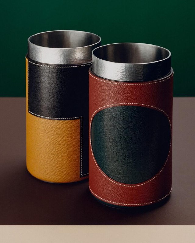 Два цилиндрических сосуда с чеканным палладием и кожаной отделкой разного цвета на тёмном фоне — предметы интерьерной коллекции Hermès.