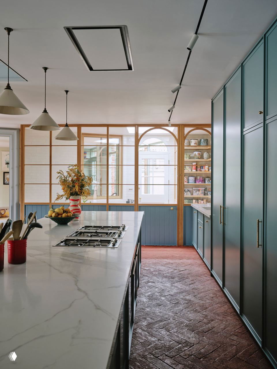 Кухня с длинным мраморным островом, купольными подвесными светильниками и голубовато-зелёными встроенными шкафами; деревянные арочные перегородки впускают свет.
