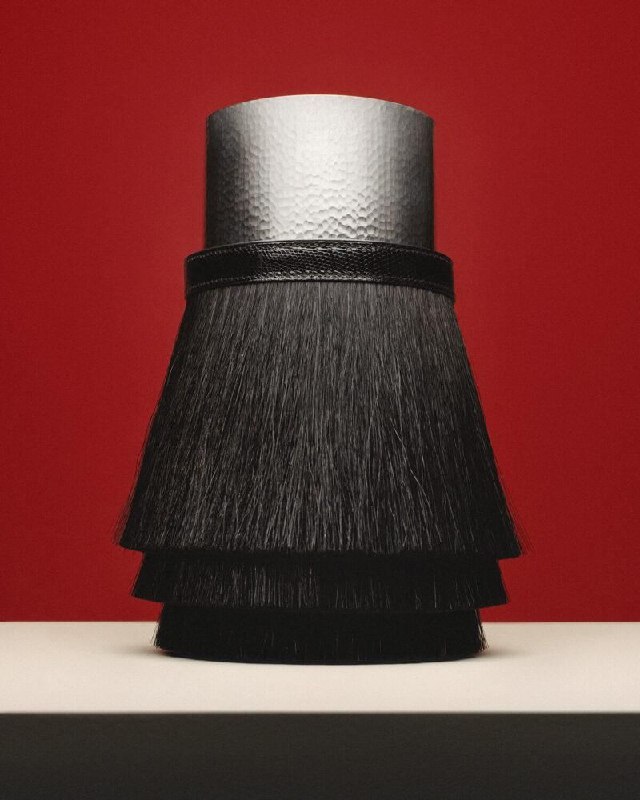 Высокий предмет с черными кистями или бахромой на фоне насыщенно-красной стены; деталь коллекции, отсылающая к конной теме Hermès.