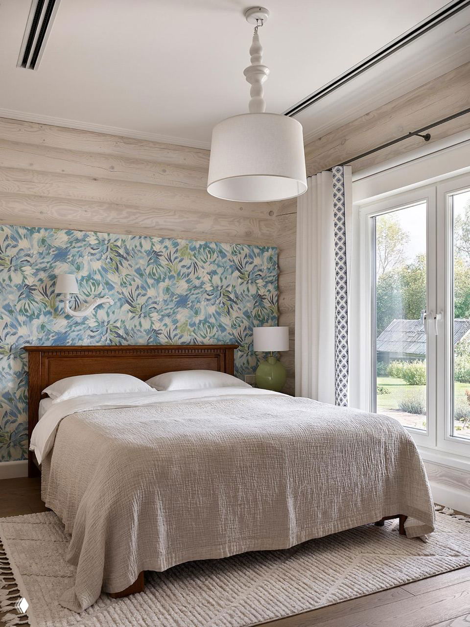 Спальня с деревянной кроватью, текстильной нейтральной постелью и цветочным растительным обоями в изголовье на фоне бревенчатых стен.