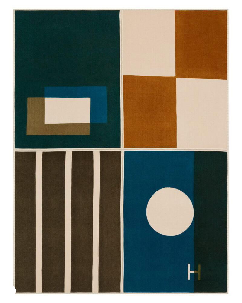 Текстильный ковёр с графическим геометрическим орнаментом в темно-зеленых, коричневых и бежевых тонах, отмеченный фирменной буквой H.