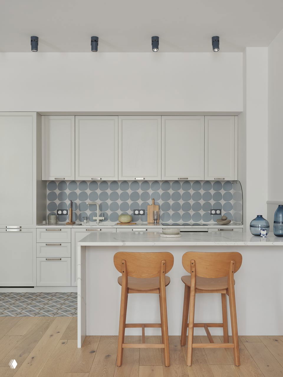 Минималистичная светлая кухня с белым полуостровом и двумя деревянными барными стульями на фоне геометрической плитки фартука и светлого деревянного пола.